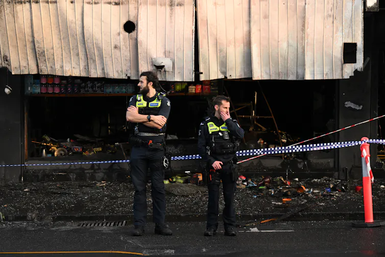 Two police officers stand in front of a burned down shop with police tape