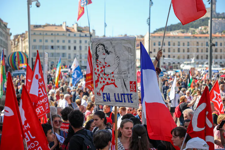 Protesters march in the streets, waving French flags and holding a labor-themed poster aloft, with French words.