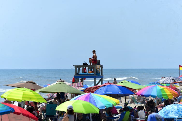 Colorful beach umbrellas cover the sand on a sunny day, with a lifeguard elevated above the crowd