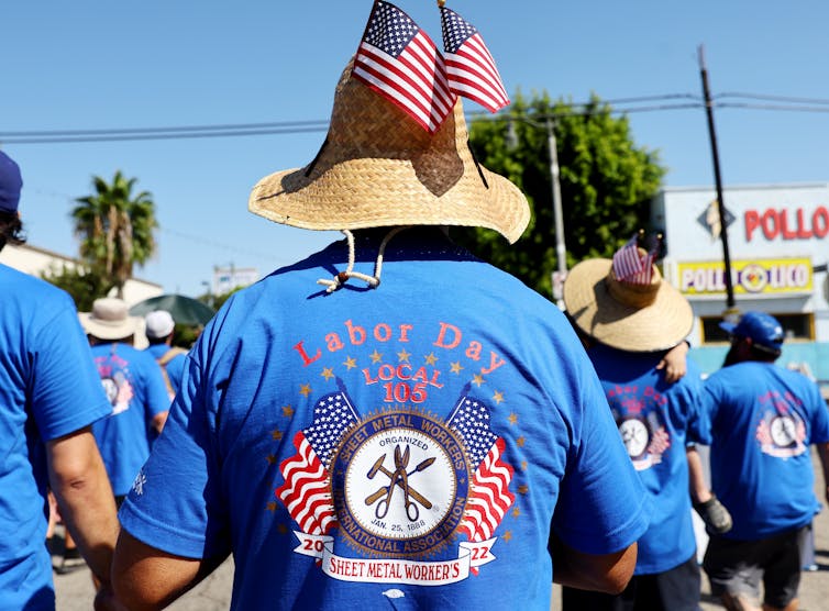 Një burrë në një T-shirt që e identifikon si anëtar të Sheet Metal Workers Local 10, duke veshur një hat prej fije me flamuj amerikanë që dalin prej tij, ecën në një paradë.