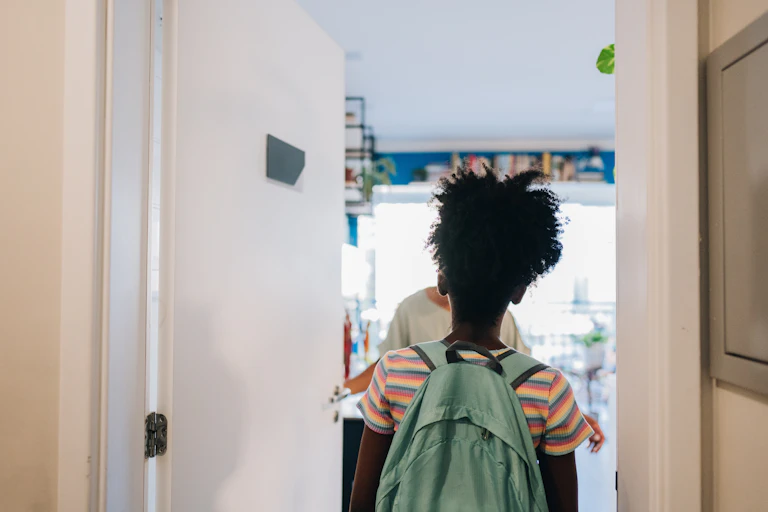 The back of a young girl wearing a backpack standing in front of an open door, which has been opened by a woman who's greeting her.