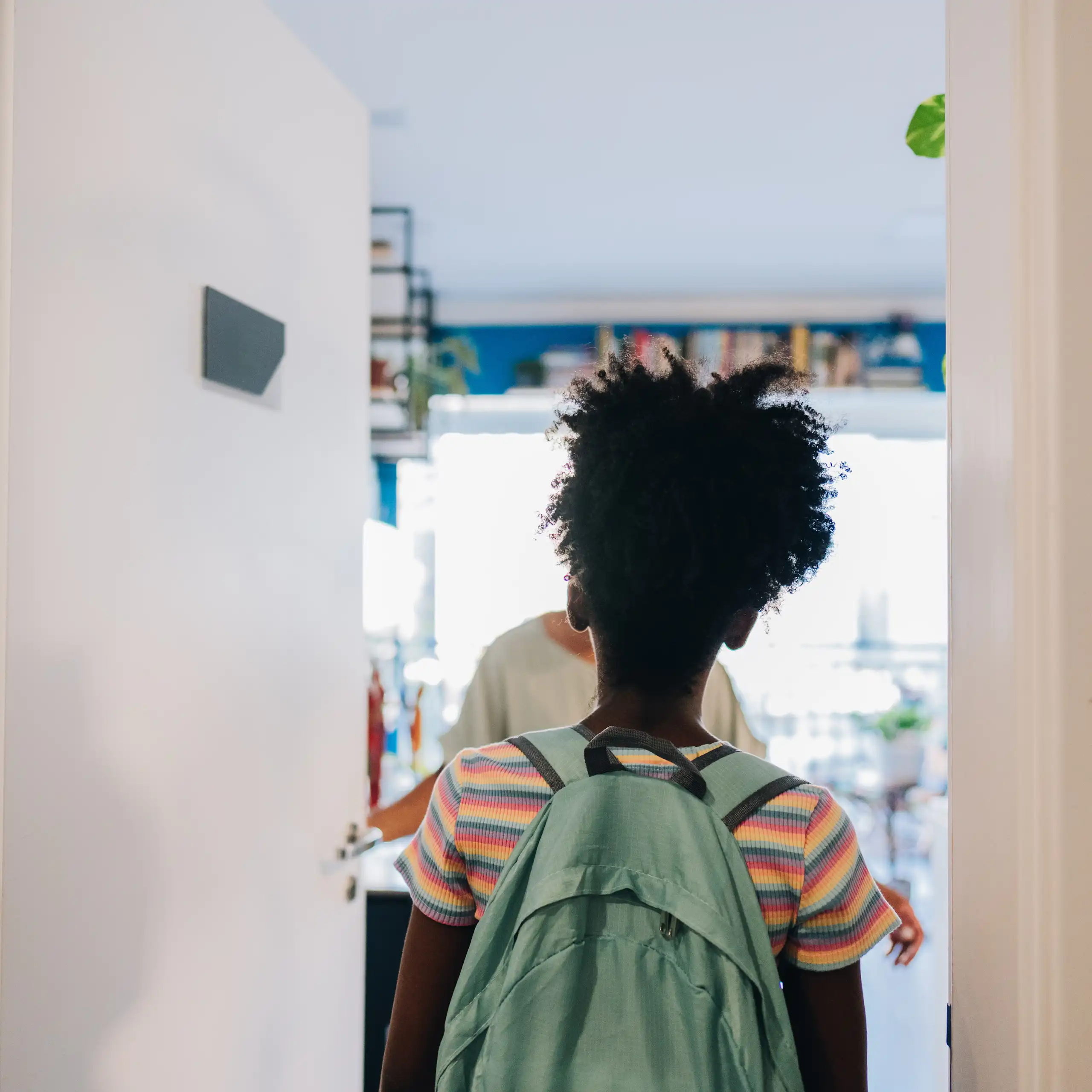 The back of a young girl wearing a backpack standing in front of an open door, which has been opened by a woman who's greeting her.