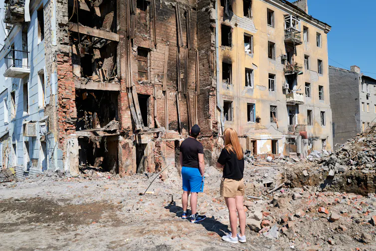 Deux personnes debout sur des décombres à côté d’immeubles de plusieurs étages bombardés