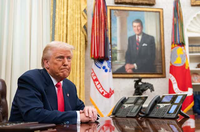 Trump at a desk, with a painting of a smiling Reagan on a wall behind him.