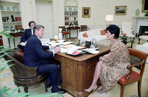 A woman sits in a chair next to the president's desk. Reagan is smiling as he talks with her.