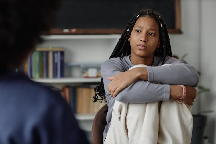 Teen girl with long braids cradles knees and listens to woman
