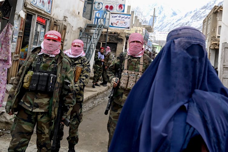 Several masked security personnel walk alongside a burqa-clad woman on a street.