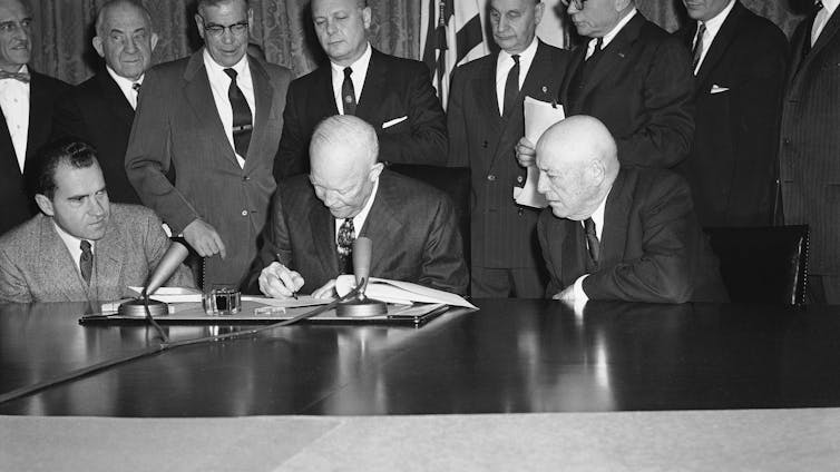 a man in a suit leans over a table to sign a document as other men in suits look on