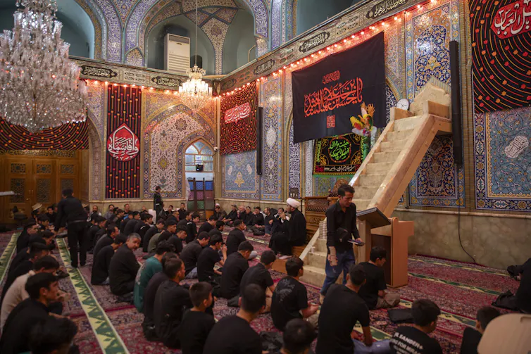 People seated in rows on a carpet, listening to a man in a black robe and white turban, in a hall with ornate decorations and a large chandelier.