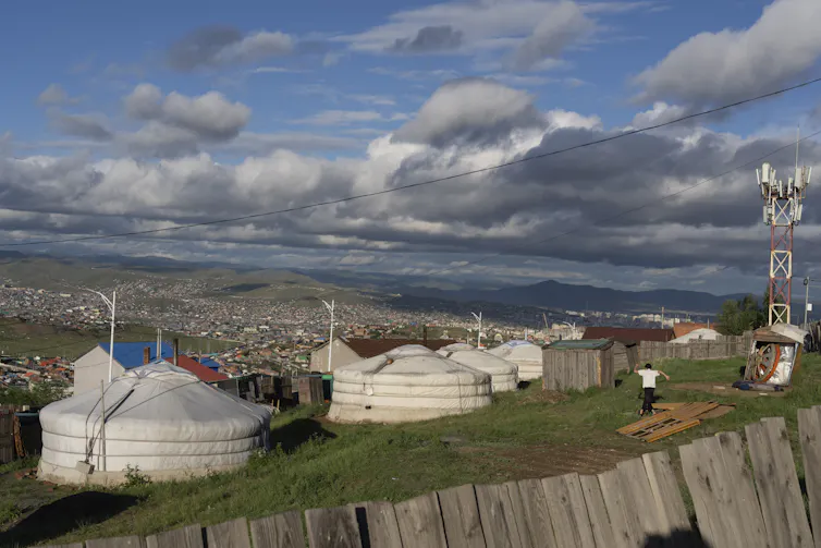 A collection of round white tents on a grassy hillside, neighbourhoods with other buildings can be seen in the background on other hillsides