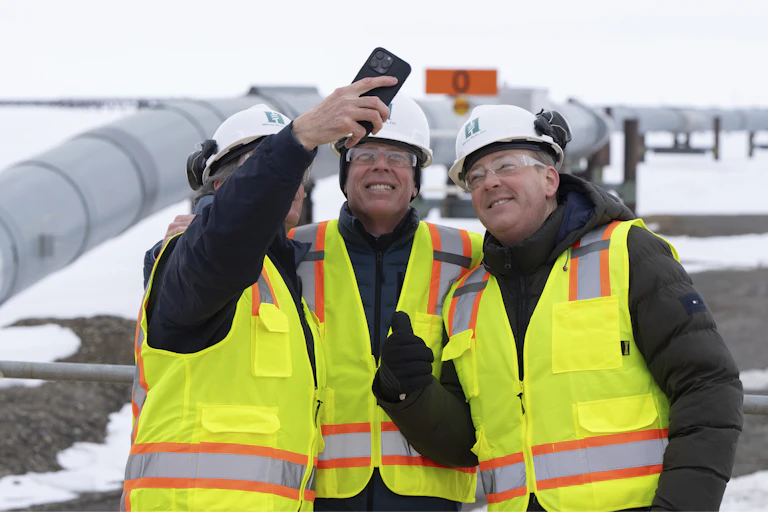 Three men in yellow safety vests take a selfie in front of a large oil pipeline.