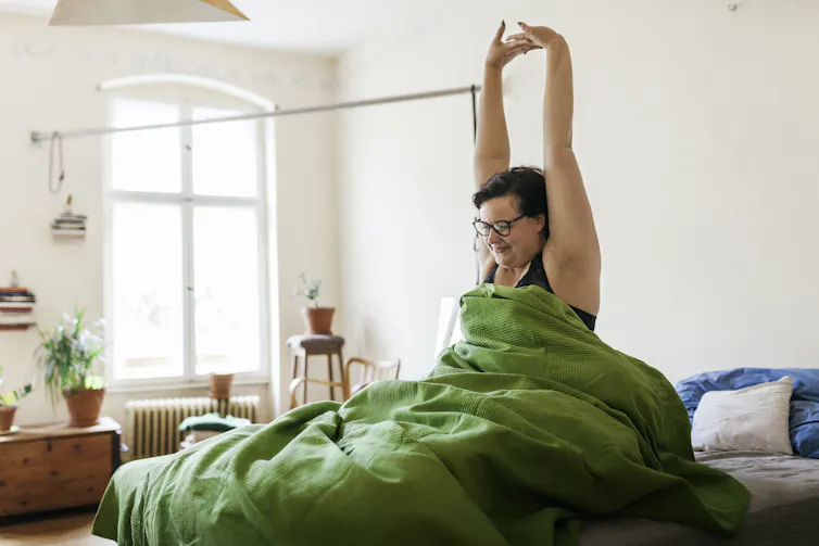 Woman sitting in bed with green eiderdown wearing black crop top stretches arms above head.