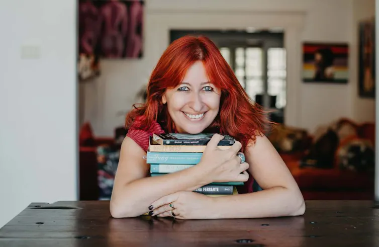 A red-haired woman holds a stack of books.