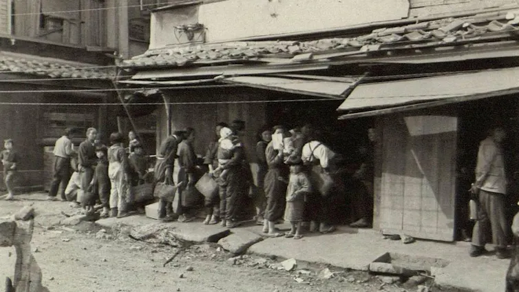 A sepia-coloured image of a group of Japanese people queueing on a street for food.