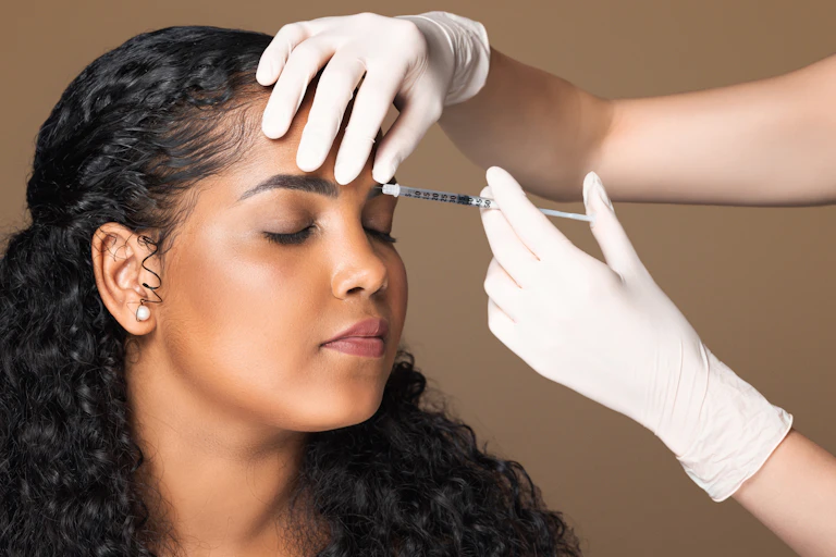 A young woman receives a botulinum toxin injection to her forehead from a woman using a white pair of surgical gloves and a needle.