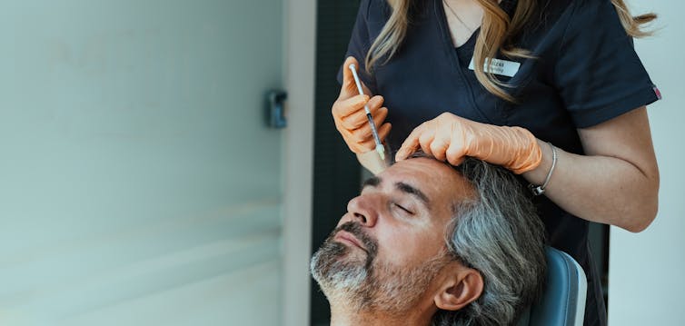 An older man receives a Botox injection into his forehead from a woman who is wearing scrubs and organ surgical gloves.