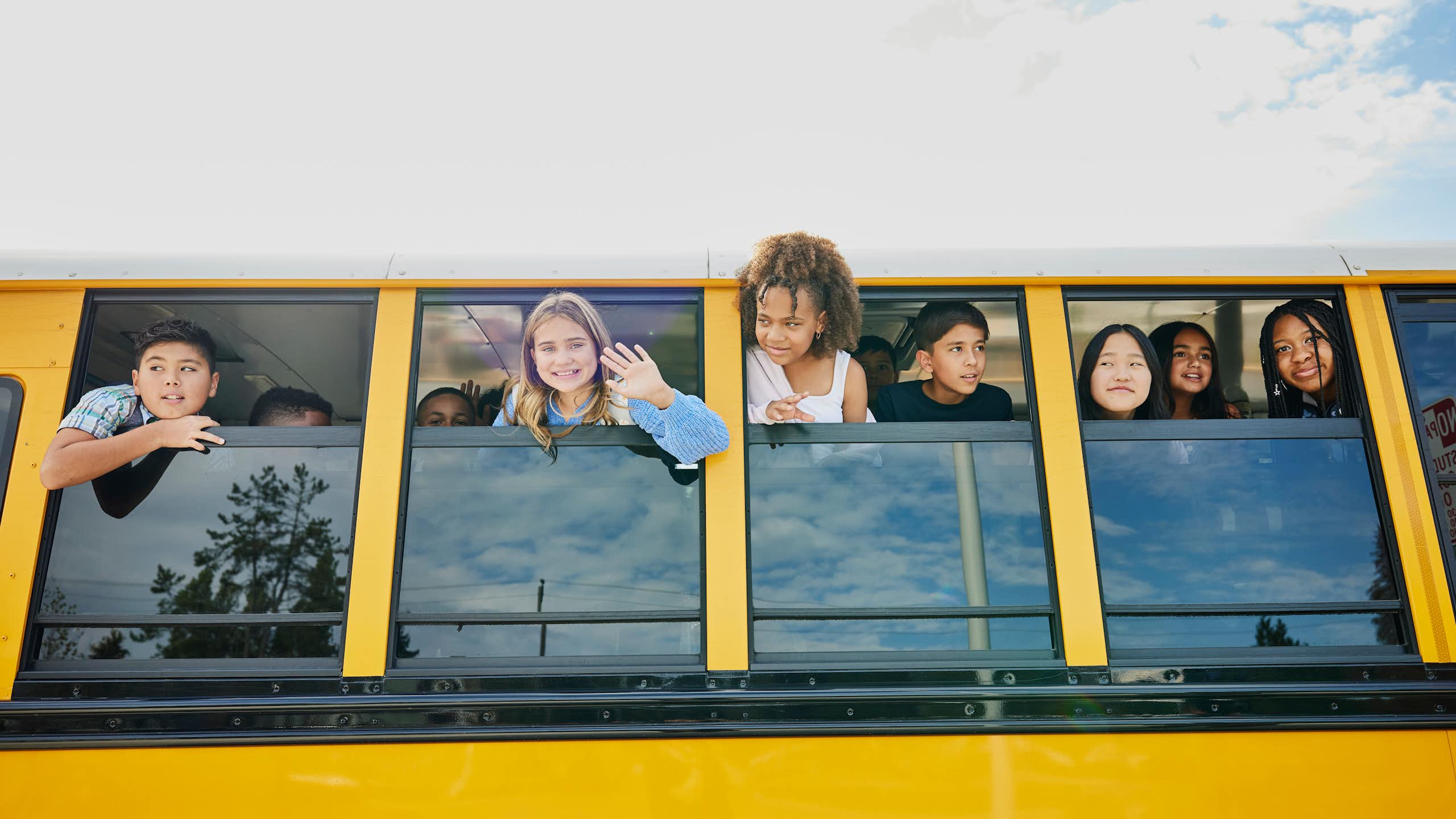 Children stick their heads out of a stopped yellow school bus and waving.
