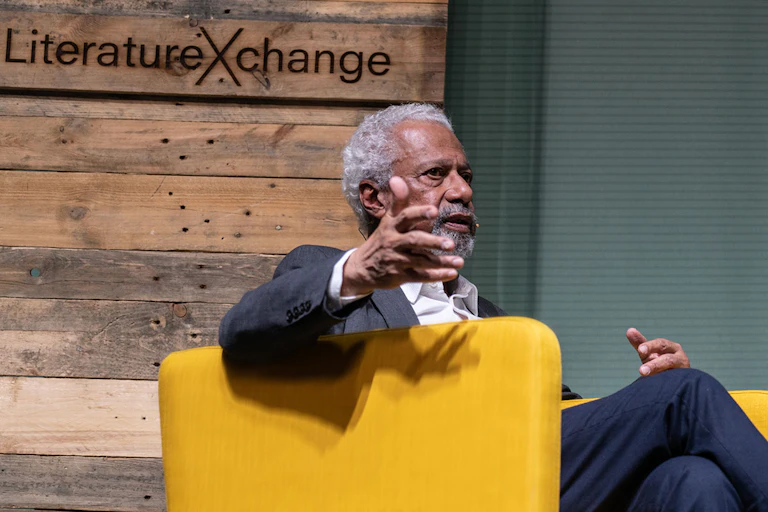 An African man with greying hair gestures as he sits in a yellow chair on a stage.
