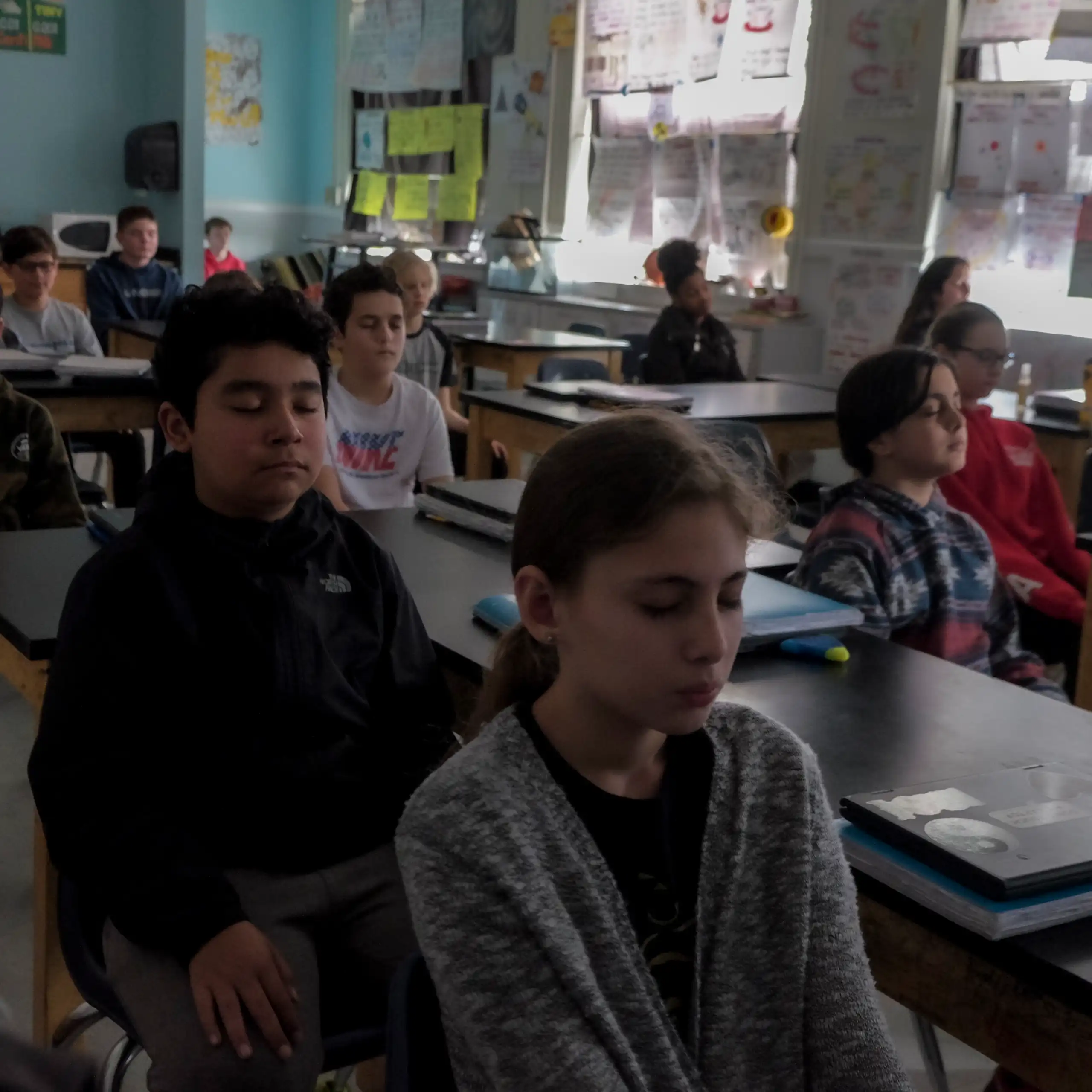 A group of children sit at individual desks and close their eyes in a classroom.