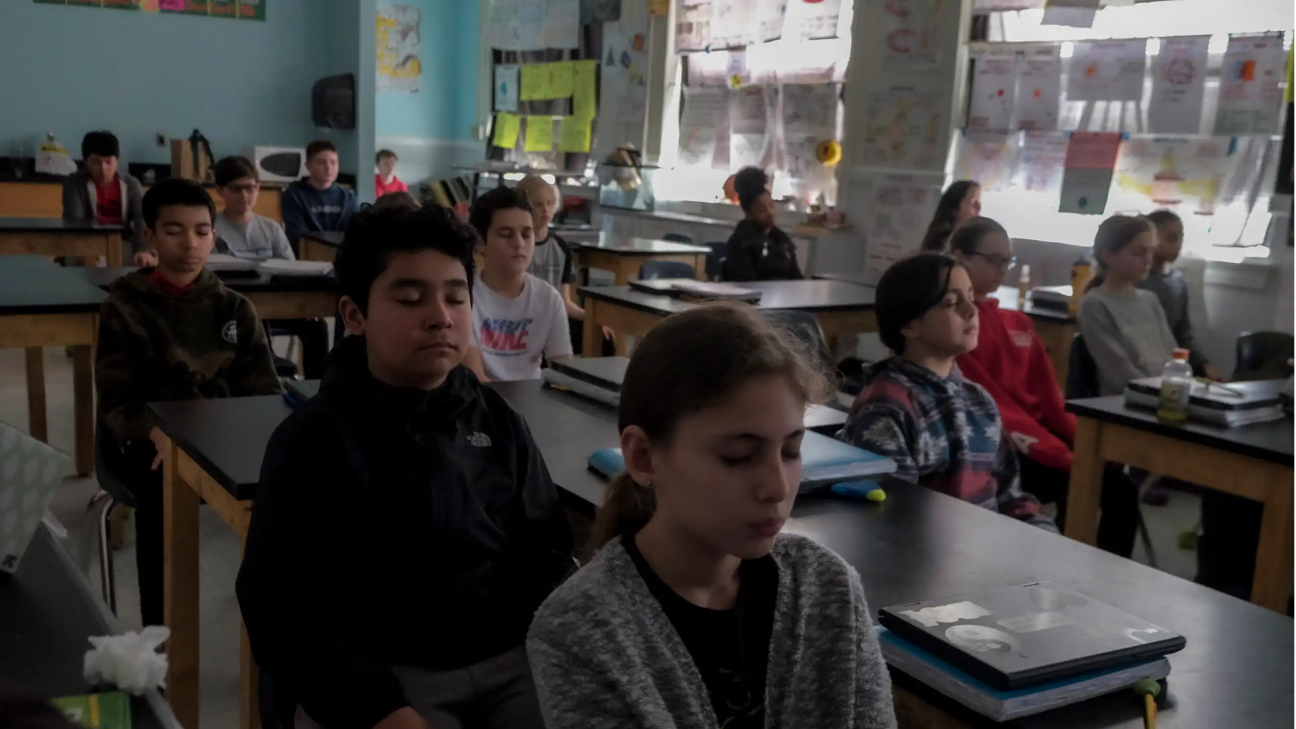 A group of children sit at individual desks and close their eyes in a classroom.