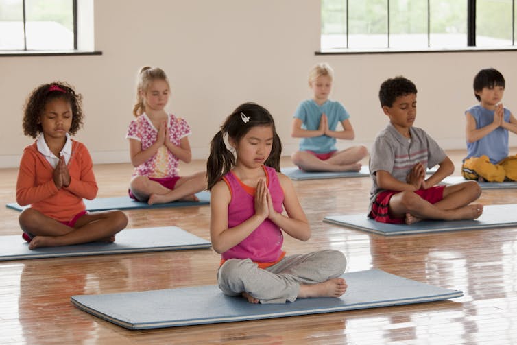 A group of young kids sit on yoga mats, close their eyes and hold their hands in a prayer position.