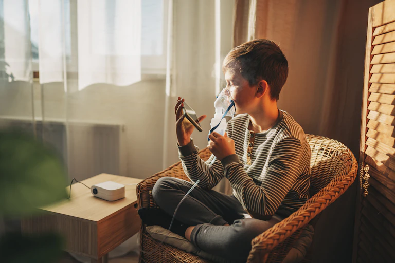 Boy wearing mask to deliver asthma medication sitting in armchair at home looking at smartphone