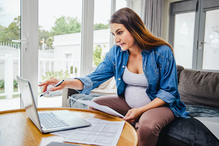 Pregnant woman sitting on sofa, holding paperwork, pointing at laptop screen