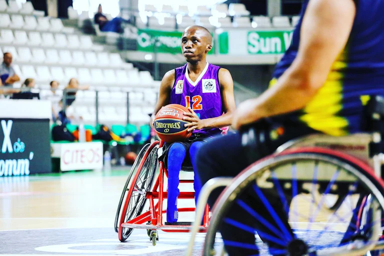 A young African man in a wheelchair holds a basketball in his lap on a court, looking ahead and about to throw it.