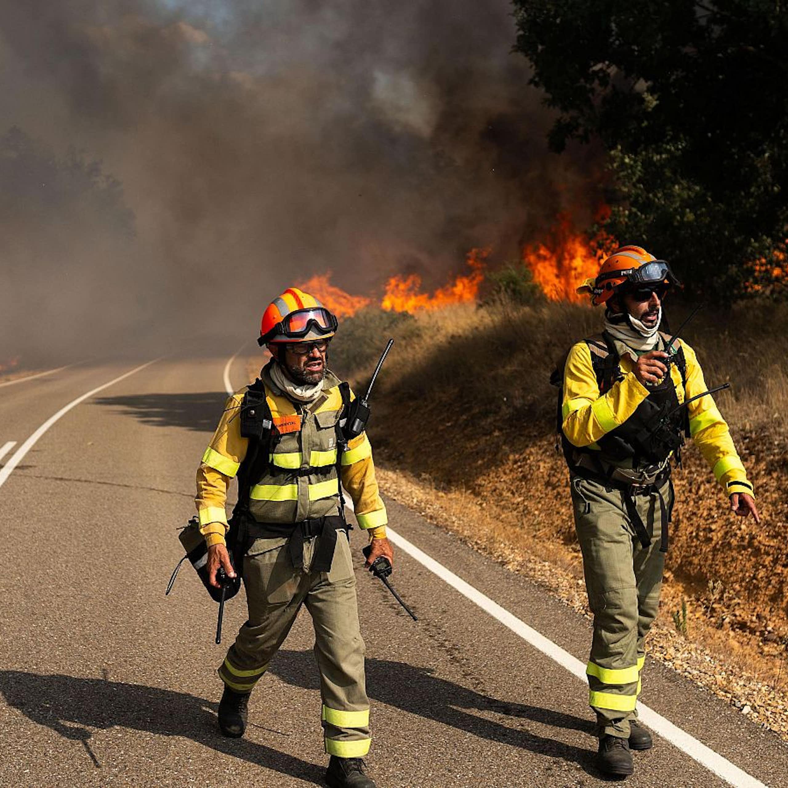 Dos hombres con trajes especiales caminan por una carretera, con llamas al fondo de la imagen