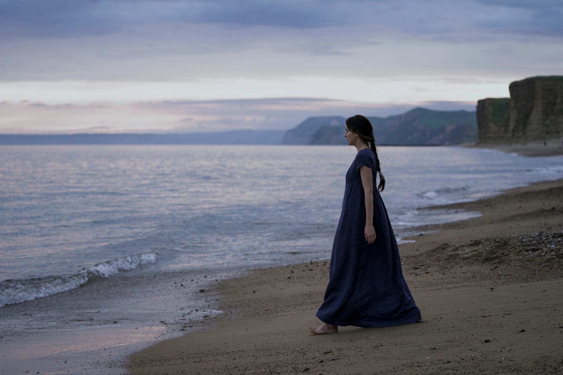 Dakota Johnson stood on a beach, in costume as Anne Elliot
