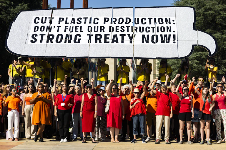 A large group of people holding a sign saying "cut plastic production don't fuel our destruction strong treaty now"