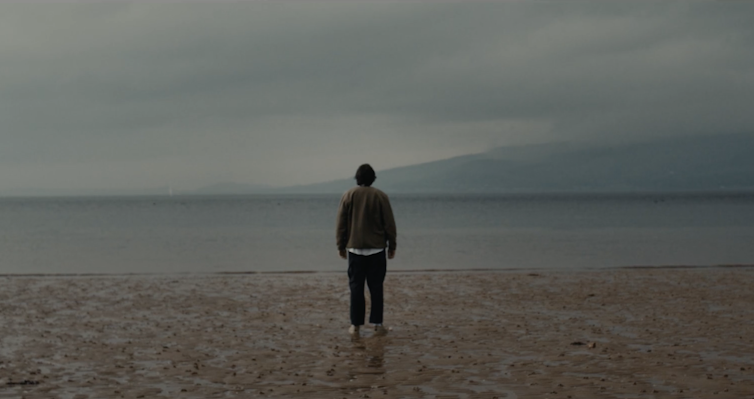A man, his back to us, stands on a foggy Scottish beach staring out to sea.