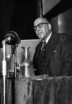 A man with glasses and wearing a suit talks from behind a table into a large microphone.