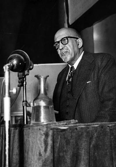 A man with glasses and wearing a suit talks from behind a table into a large microphone.