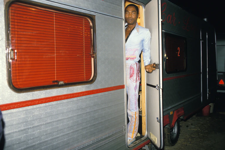 An African man in a jumpsuit peers from a caravan door that he holds ajar.