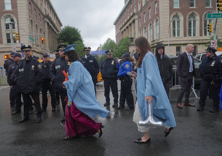 Two young women wearing long light blue graduation robes walk past a row of police officers outside two large buildings on a gray day.