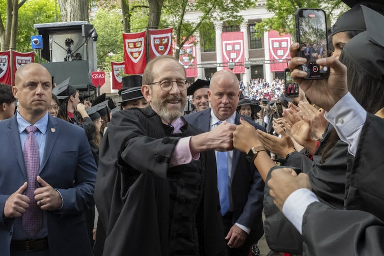 An older white man with a beard, flanked by two men in suits, bumps fists with a young person in a crowd.