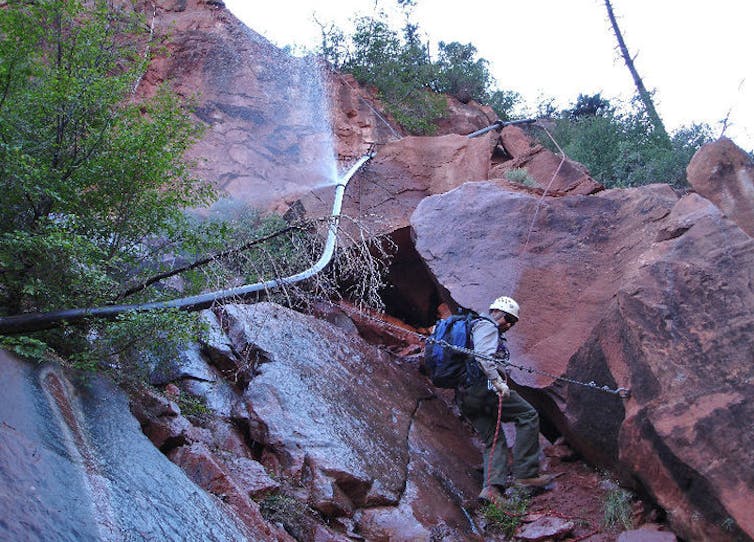 A worker in a hard hat picks his way carefully over wet rocks below a split in an exposed water line.