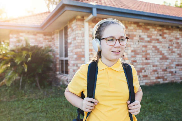 A young girl in headphones and a school uniform stands in a front yard with a back pack.