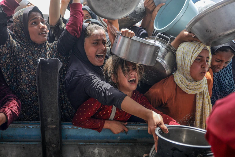 Des femmes et des fillettes attendent pour recevoir un repas, elles crient et tendent des contenants alimentaires type casseroles et bassines en métal et en plastique.