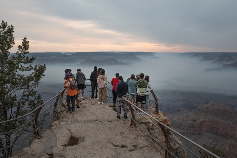 People stand on the canyon's rim behind a rail. A layer of smoke fills the canyon.