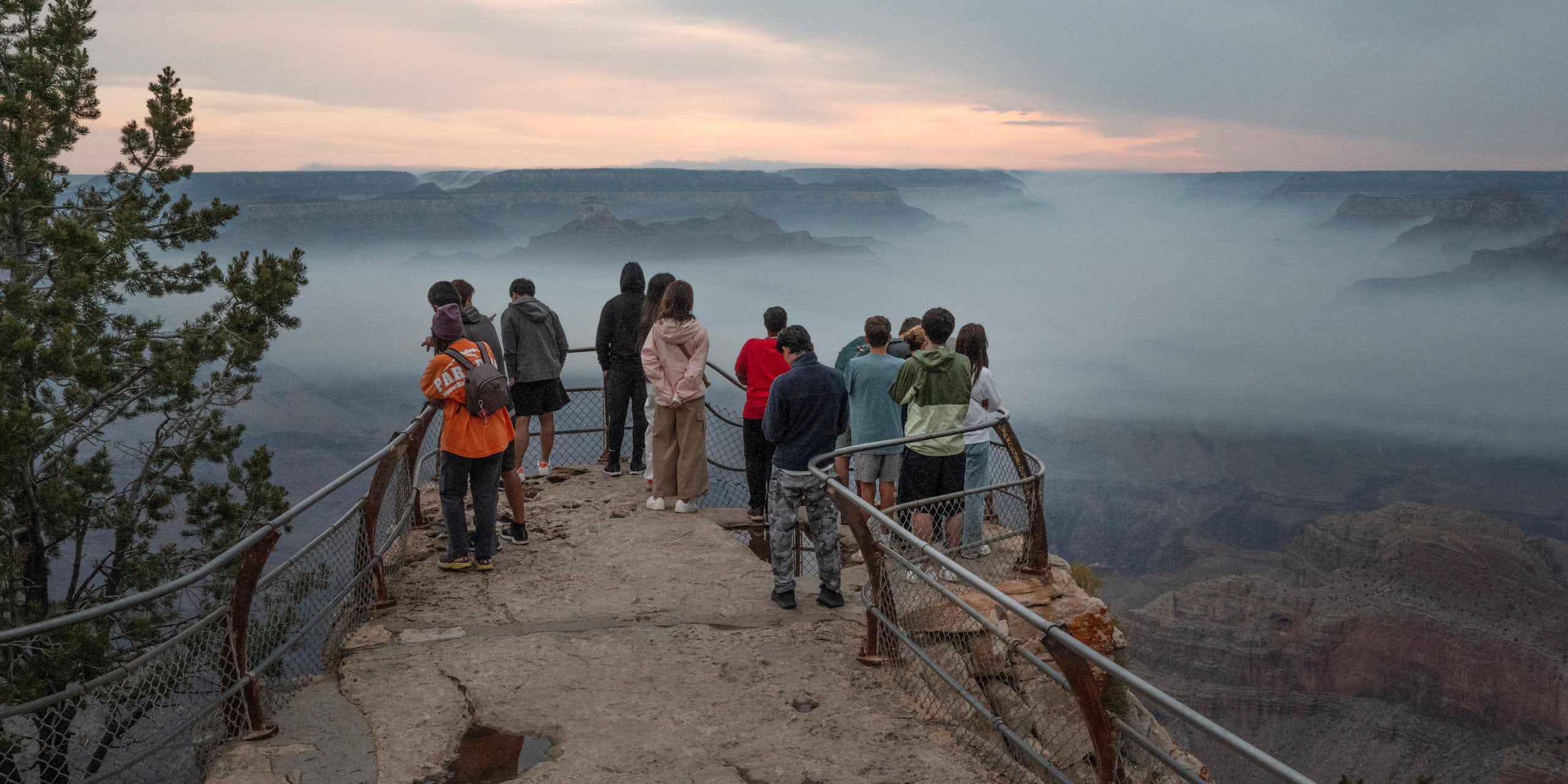 People stand on the canyon's rim behind a rail. A layer of smoke fills the canyon.