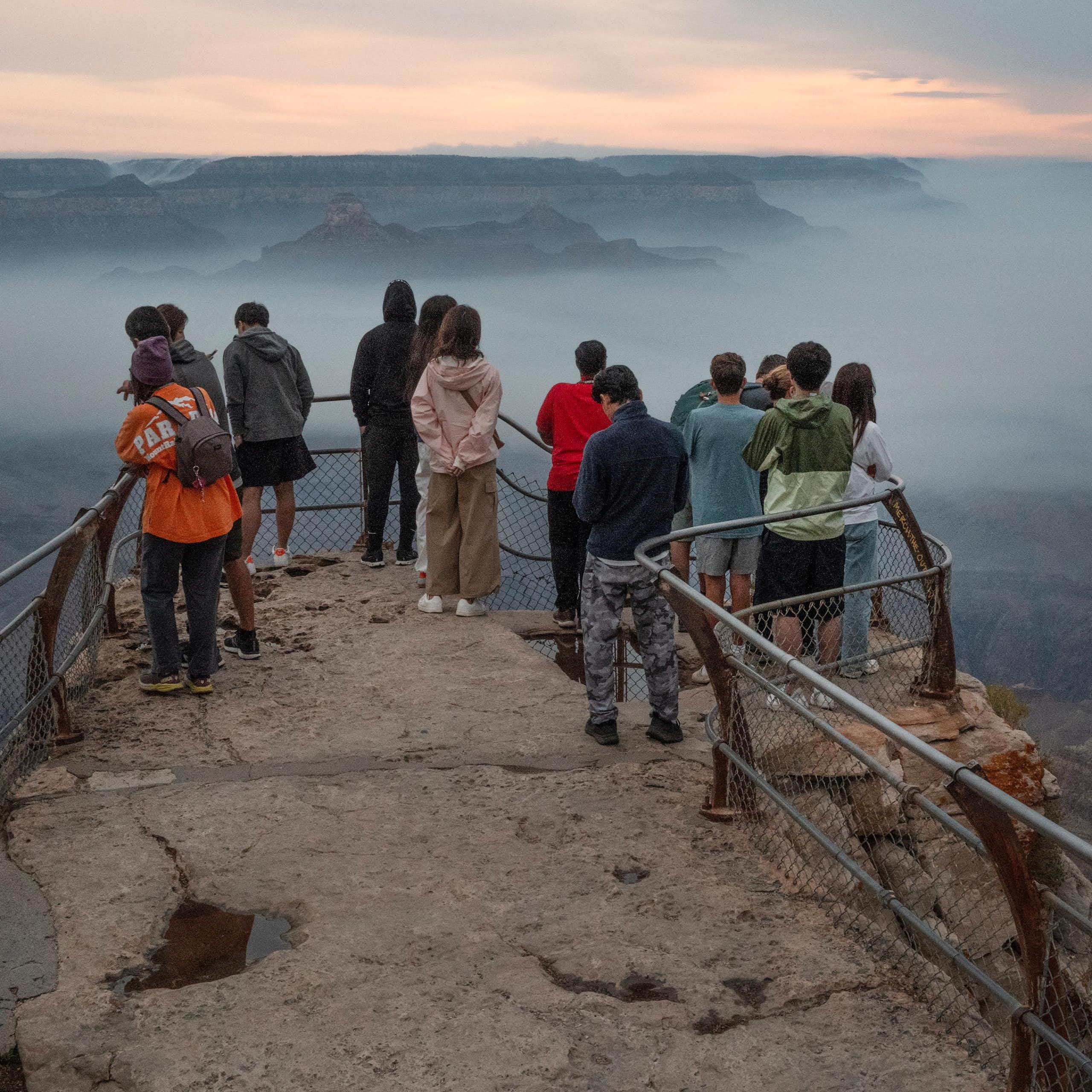 People stand on the canyon's rim behind a rail. A layer of smoke fills the canyon.