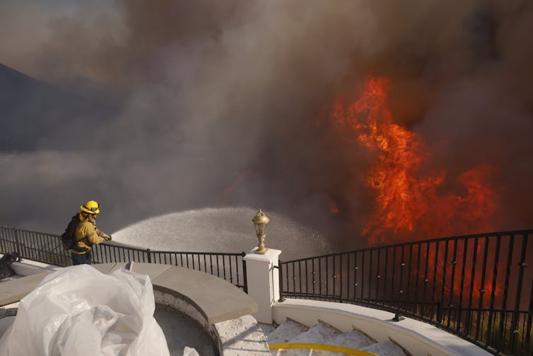 A firefighter sprays water from a hose on flames in a canyon area below a porch.