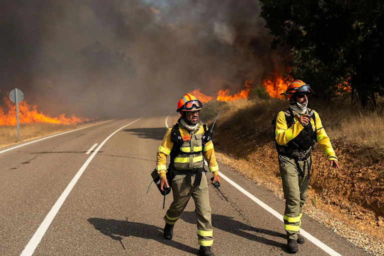 Two firefighters wearing specialised clothing walk on a road, with flames and smoke in the background.