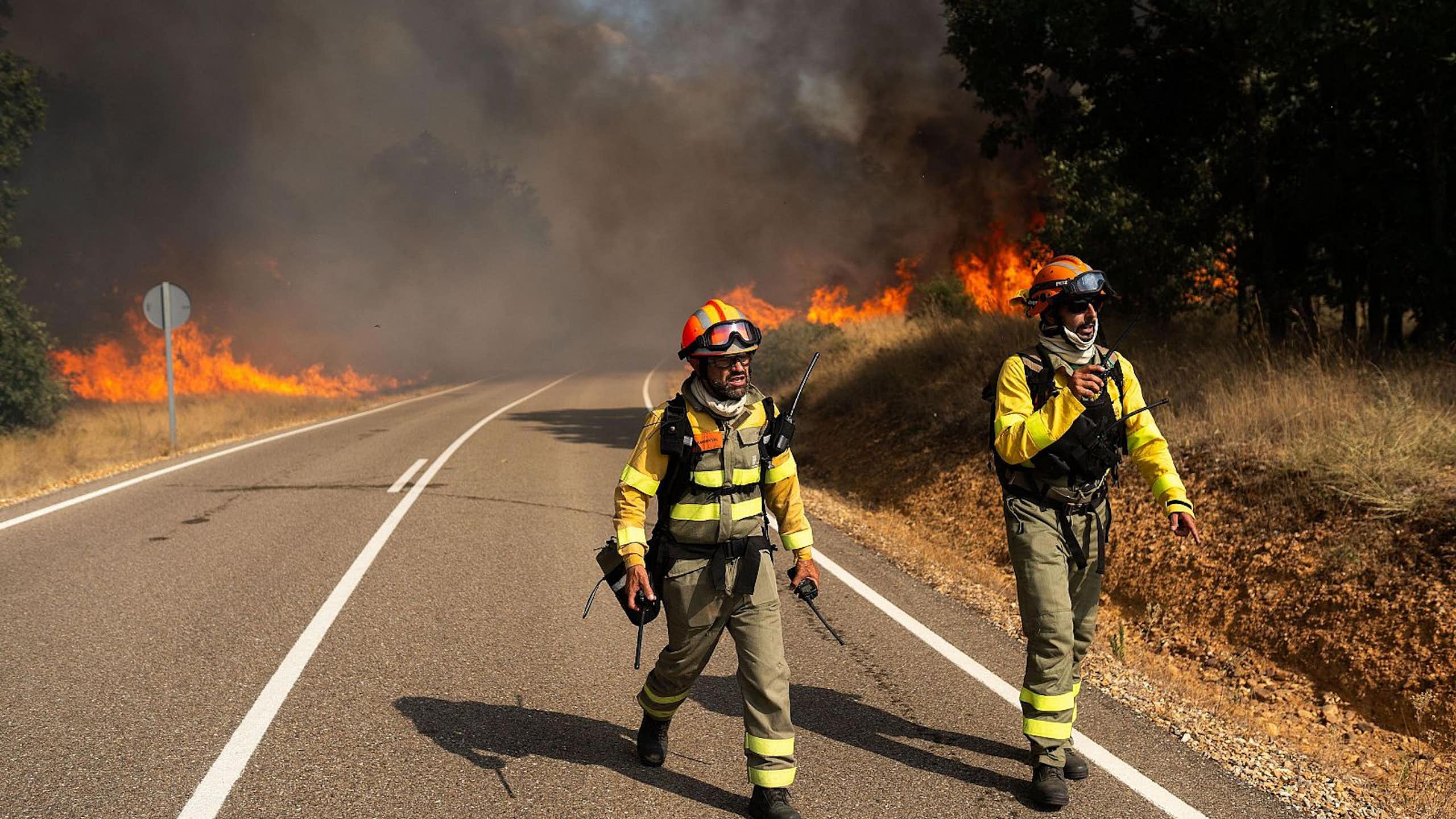 Two firefighters wearing specialised clothing walk on a road, with flames and smoke in the background.