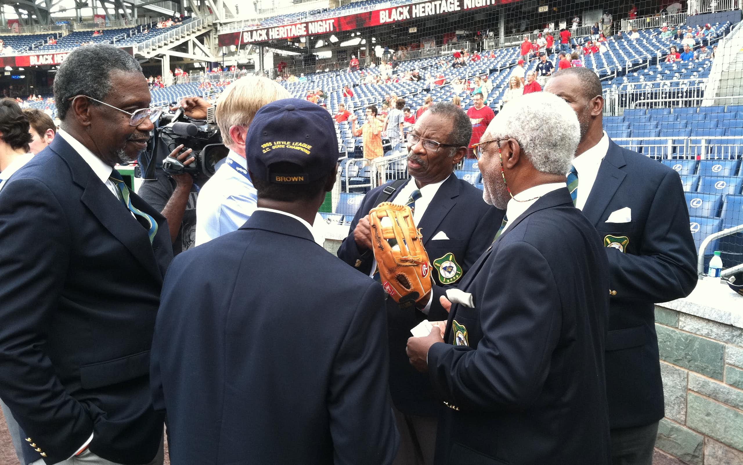 A group of older Black men wearing suits chat on the field of a baseball stadium.