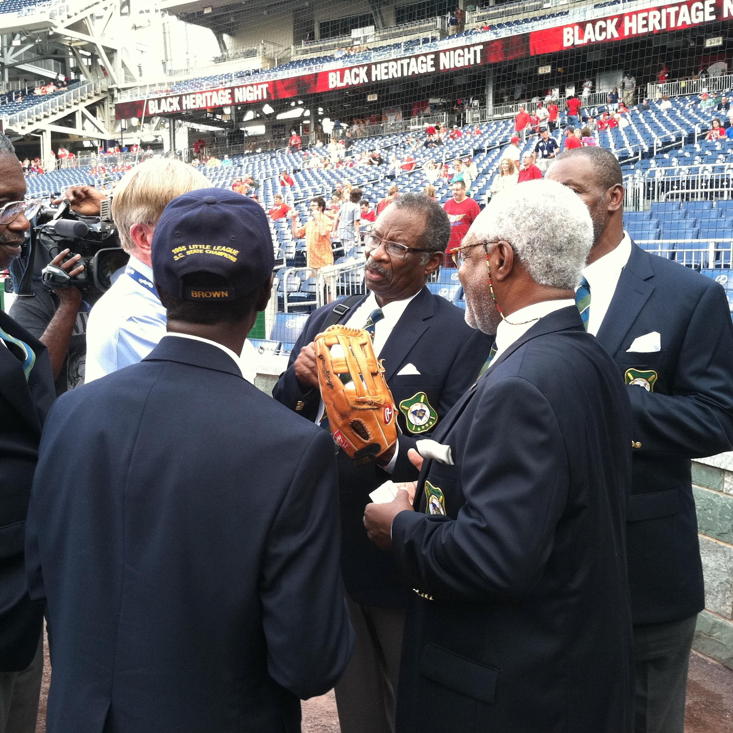 A group of older Black men wearing suits chat on the field of a baseball stadium.