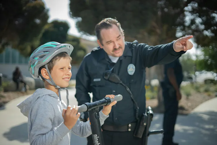 a boy on a scooter stands next to a police officer pointing