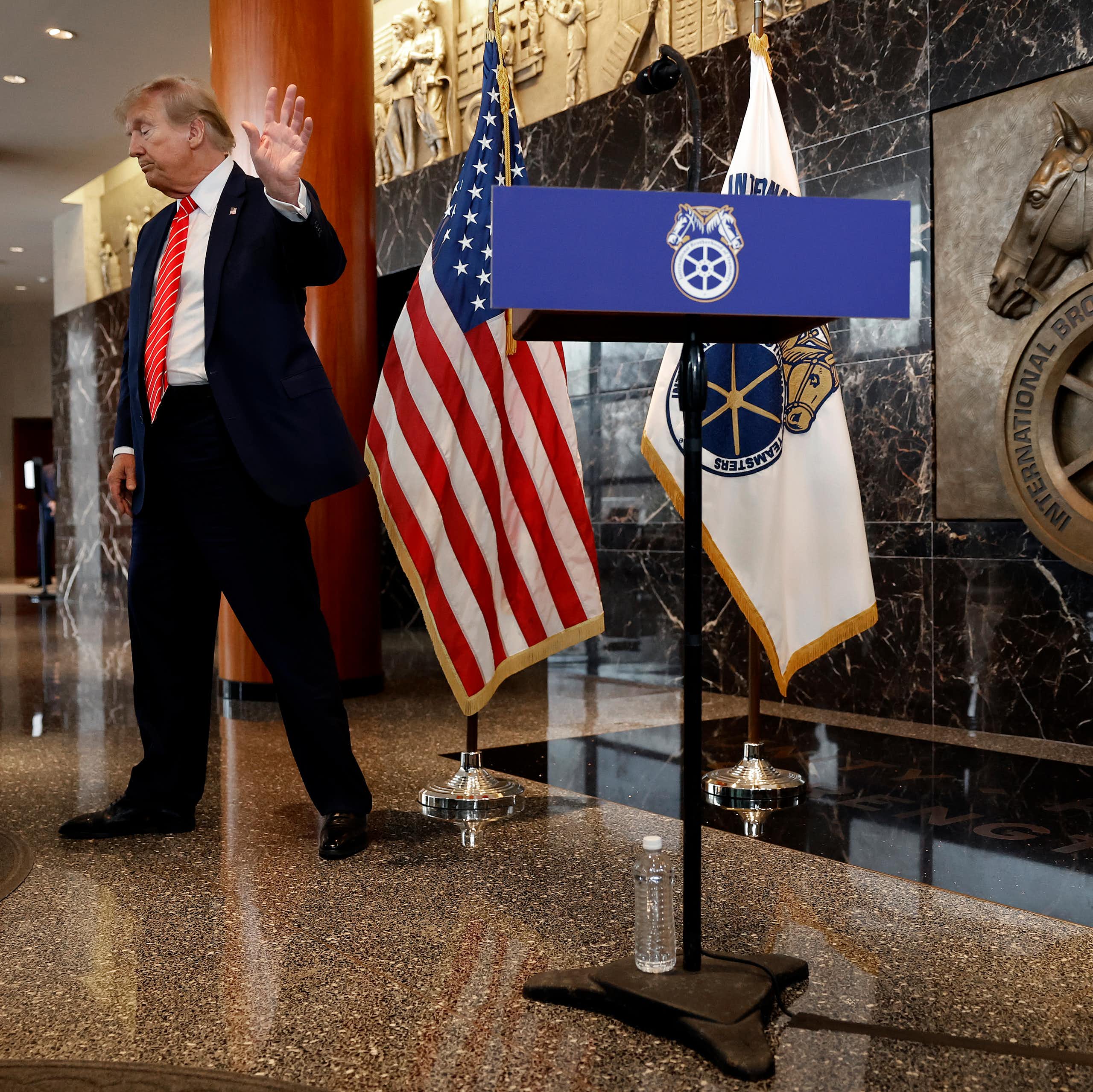 Donald Trump unenthusiastically waves goodbye while standing in a large building lobby that includes the Teamsters logo.